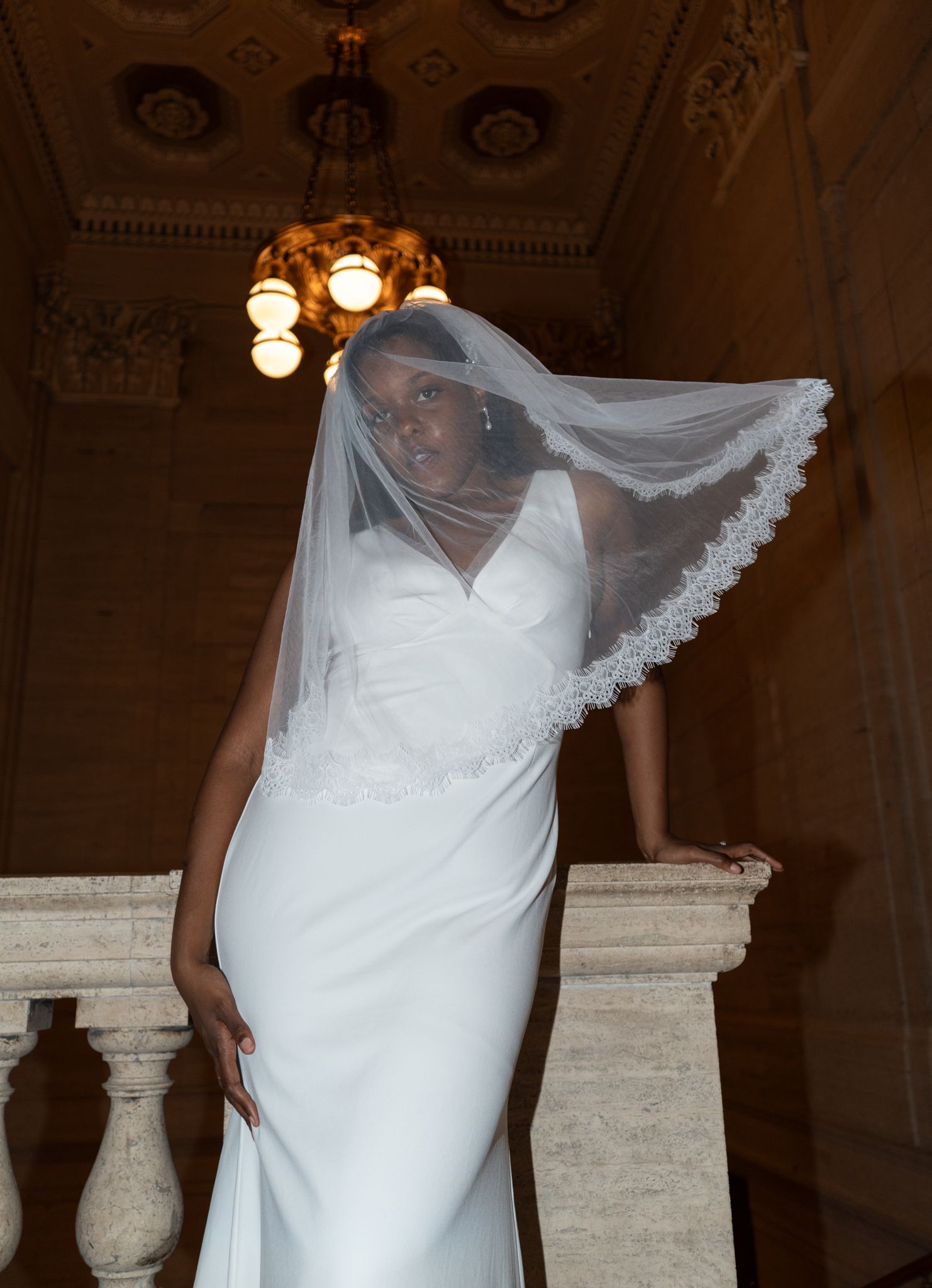Elegant Black bride in white fitted gown and lace-trimmed veil poses on marble staircase in ornate venue.