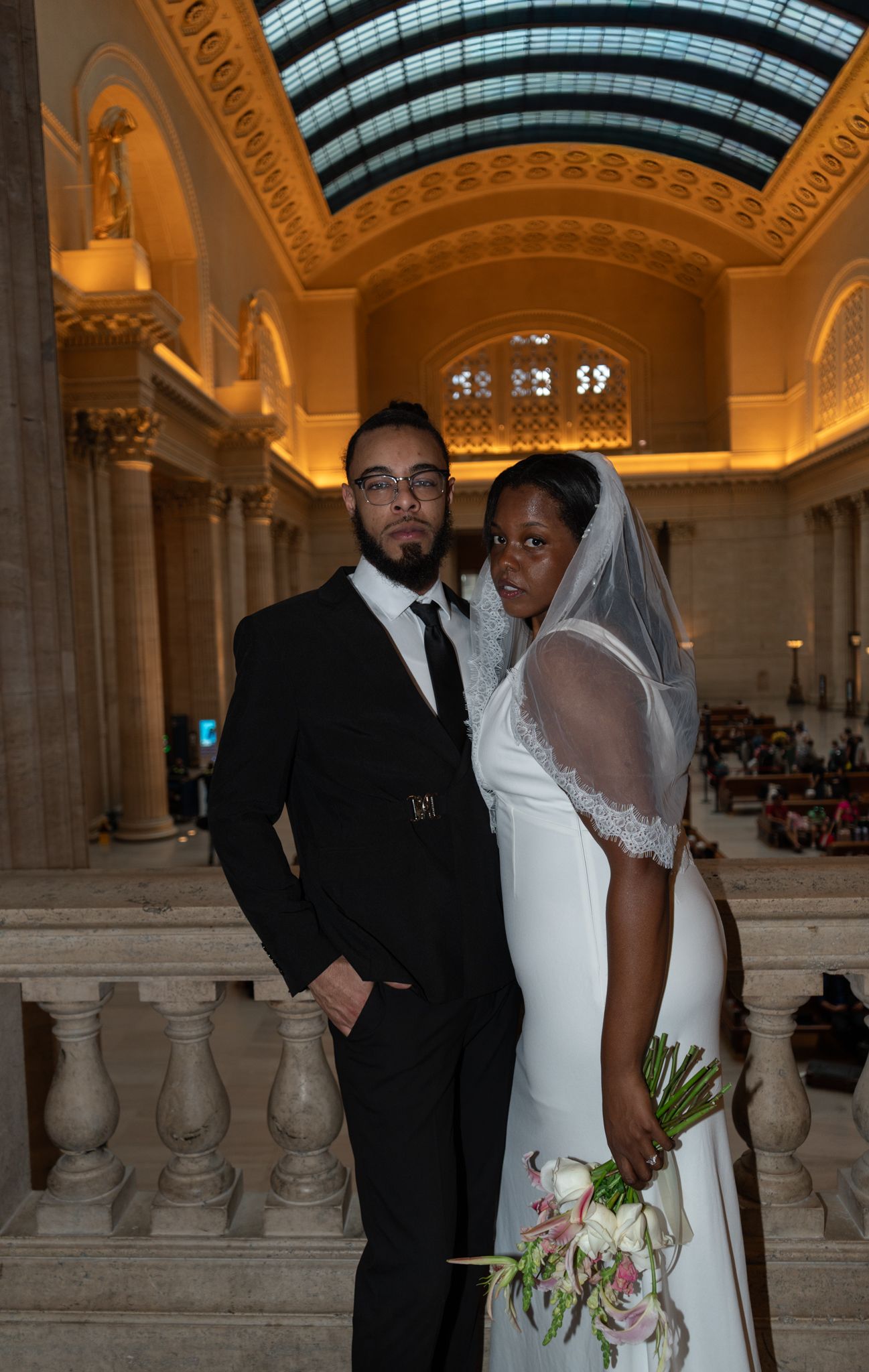 Newlywed couple posing in elegant wedding attire inside a grand hall with ornate columns and glass ceiling.