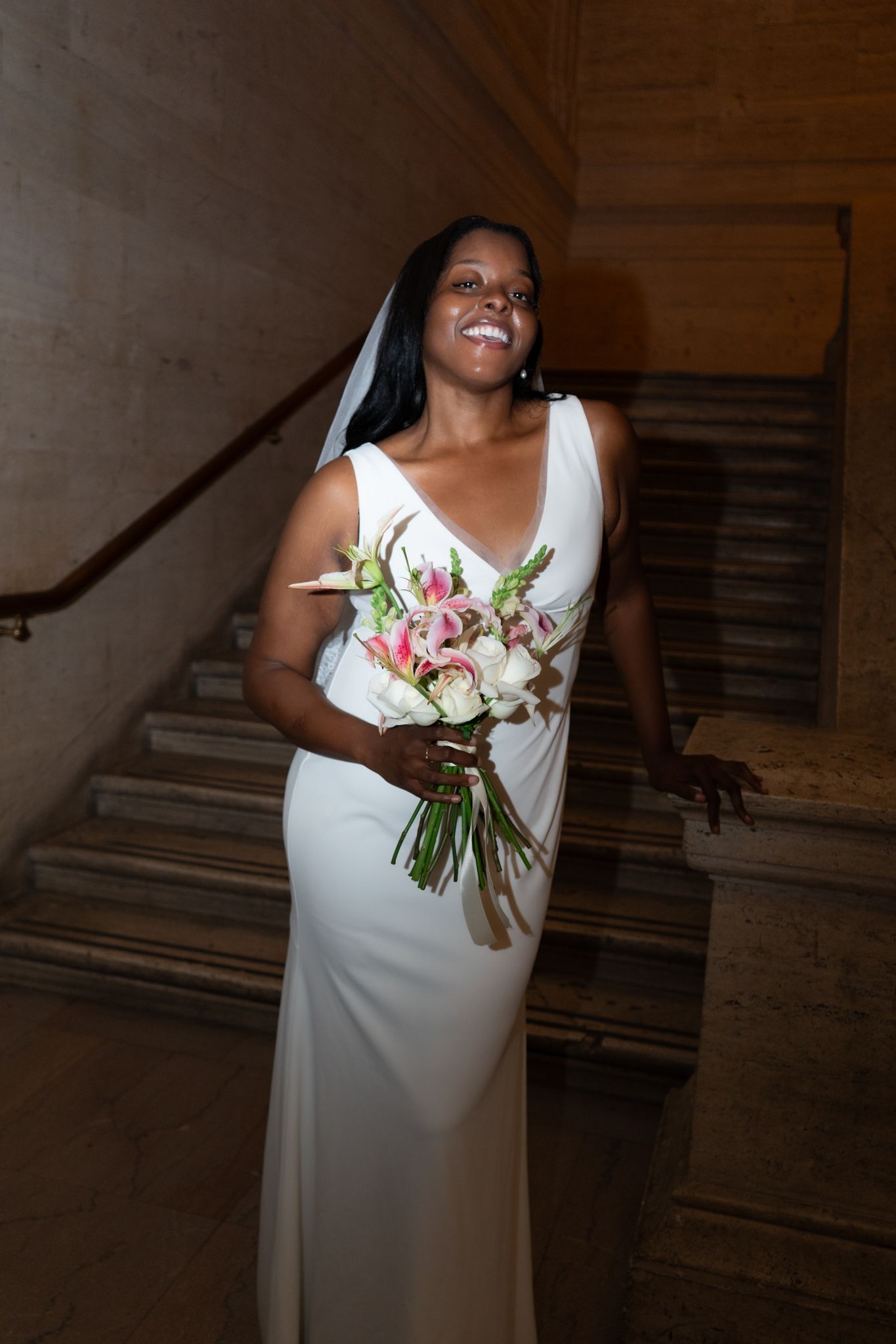 Smiling bride in white gown holding pink bouquet on marble staircase.