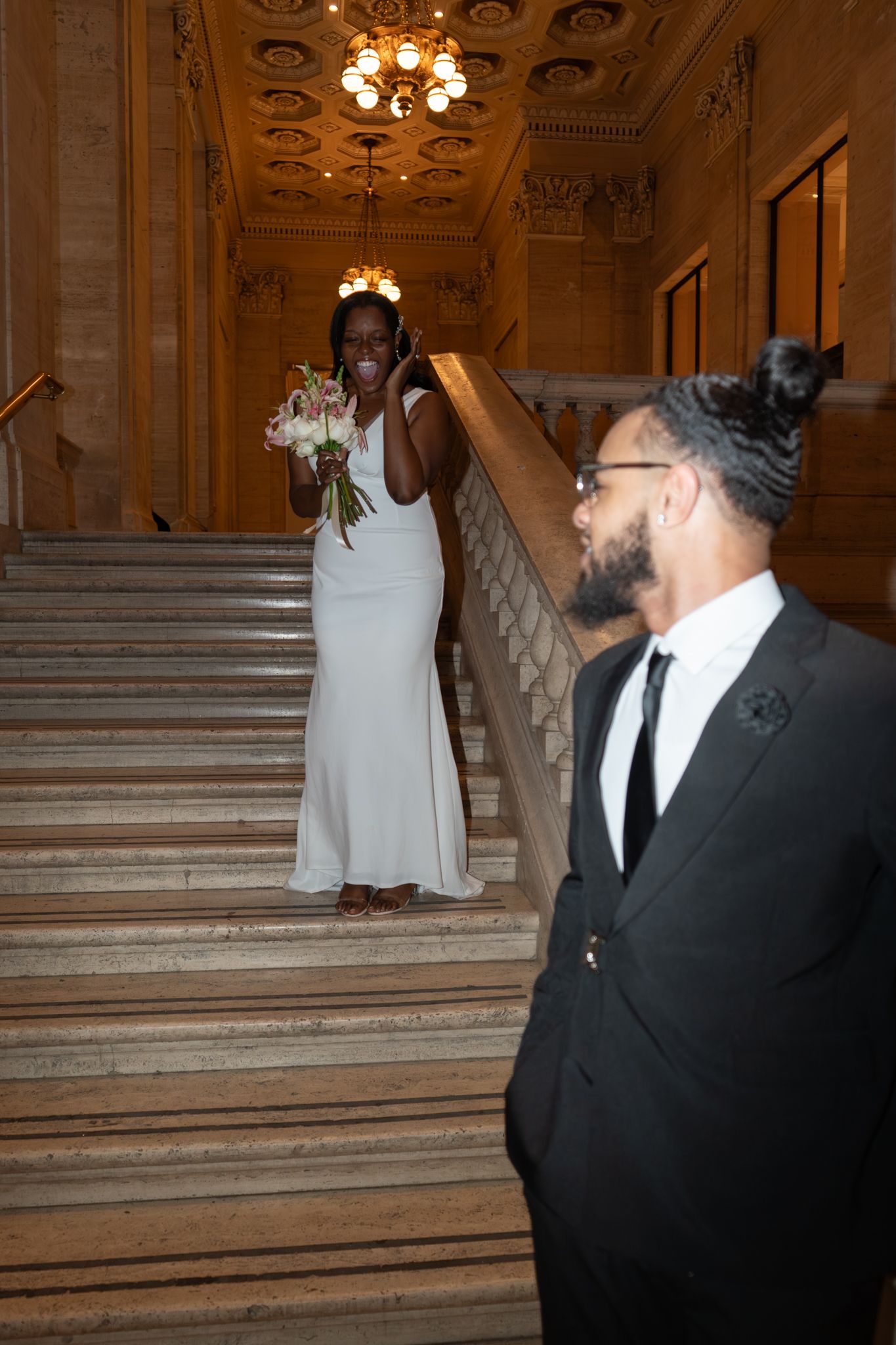 Bride in white gown holding bouquet smiles at groom in black suit on ornate marble staircase.