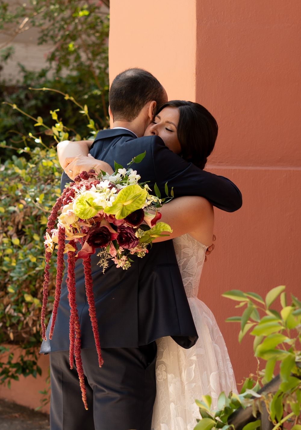 Bride and groom share a heartfelt embrace on their wedding day, bride holding a vibrant bouquet of red and green flowers.