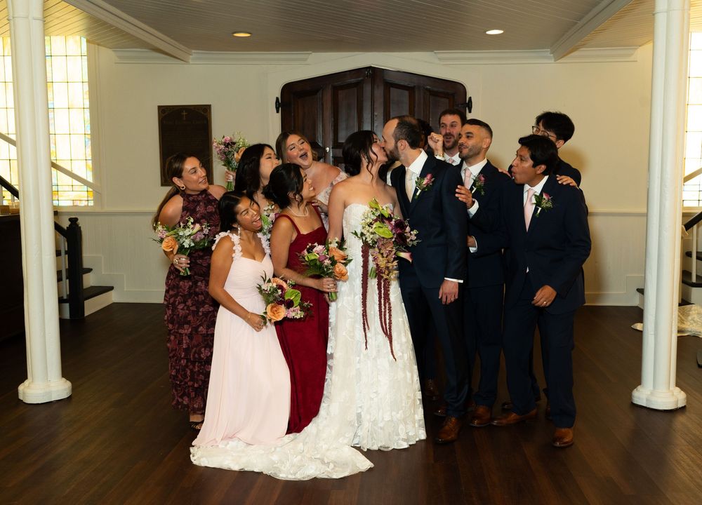 Wedding party in navy suits and burgundy dresses surrounds two brides sharing a kiss in an elegant white venue.