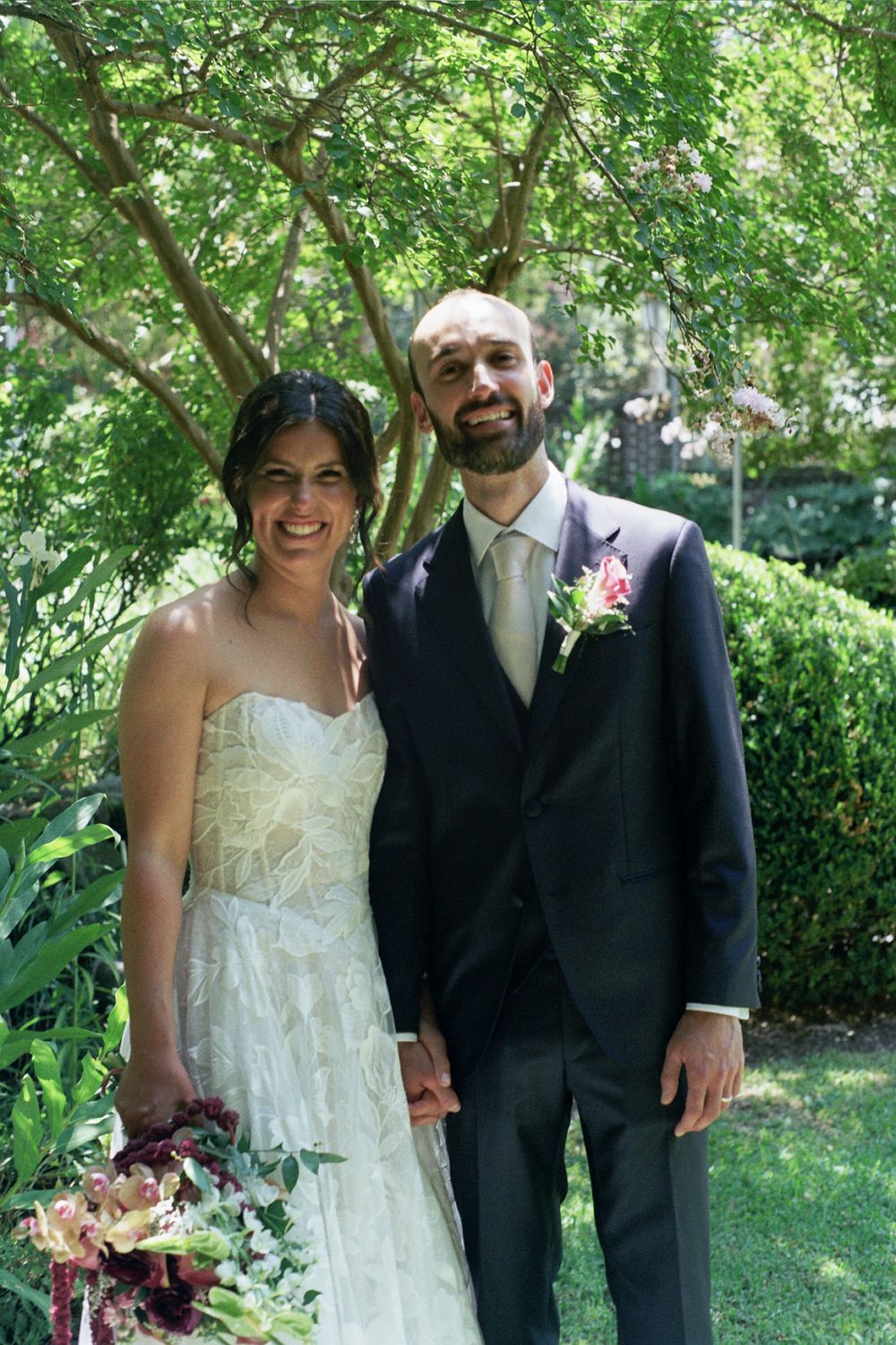 Smiling bride and groom pose together outdoors in a lush garden on their wedding day.