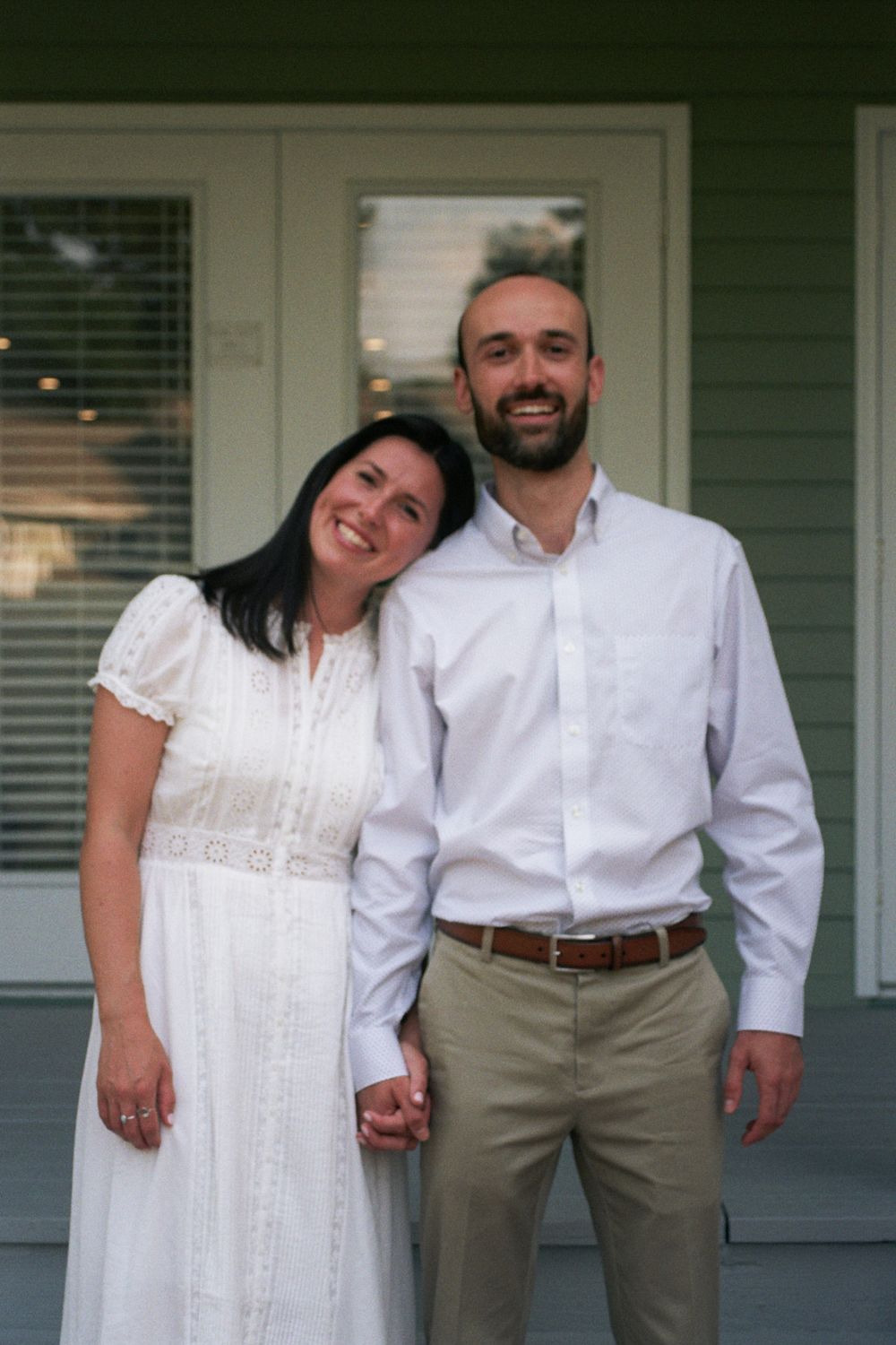 A smiling couple in white outfits pose together on a porch, the woman in a lace dress and man in a button-up shirt.