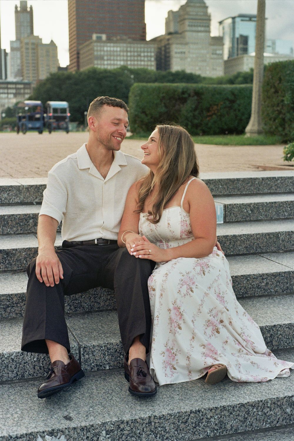 Couple sitting on stone steps smiling at each other with city skyline in background, engagement photo.