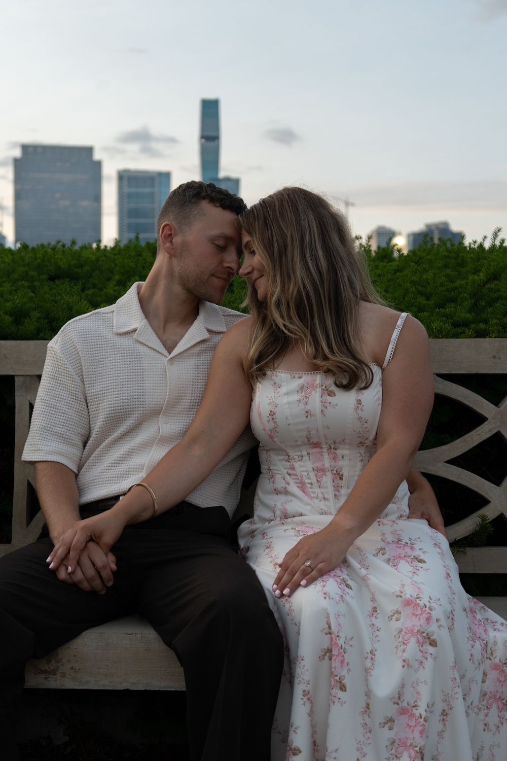 Couple sitting on a bench sharing a romantic moment with city skyline and greenery in the background.