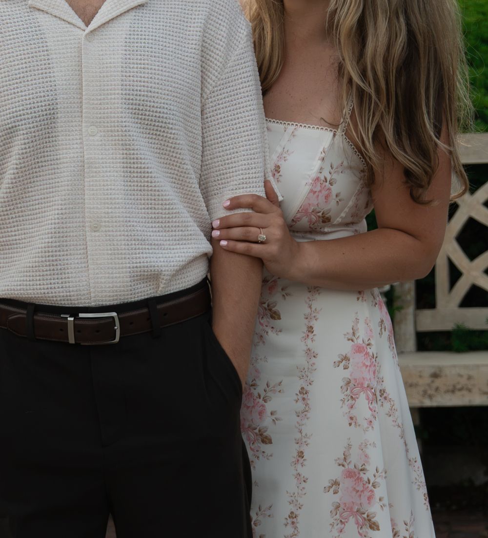 Couple posing outdoors, woman in floral dress holding mans arm, engagement ring visible on her hand.
