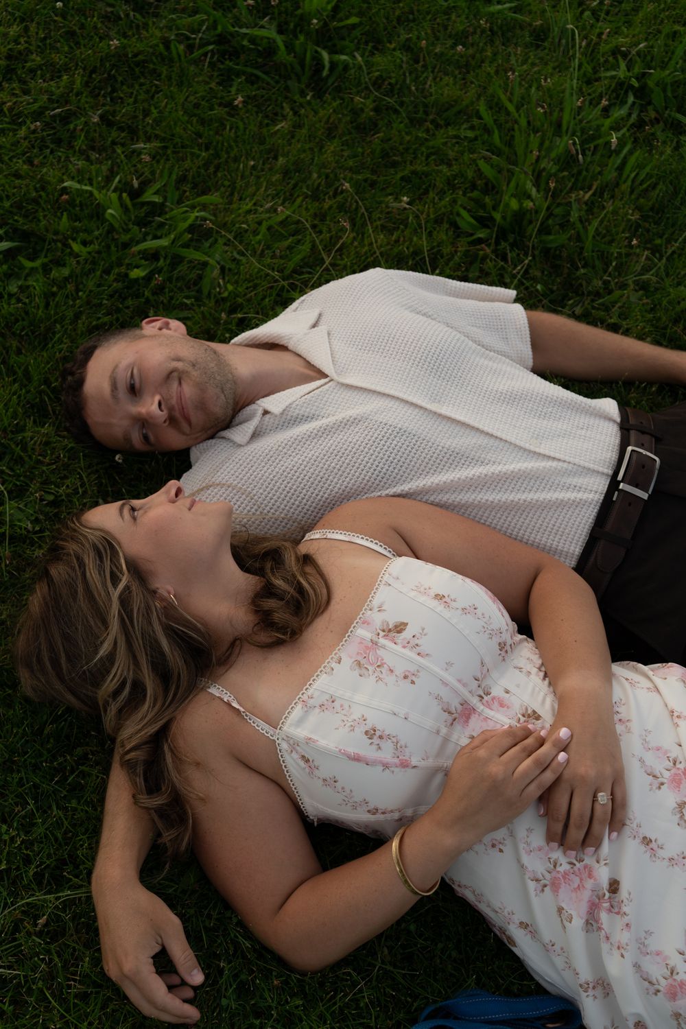 Couple lying on green grass together, woman in floral dress resting on mans chest smiling.