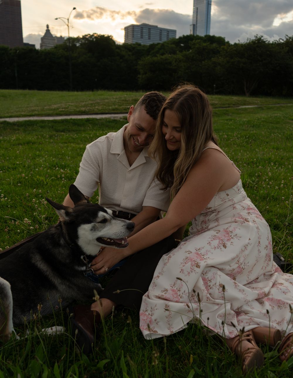 A couple sits on green grass in a park, petting their Siberian Husky dog, with a city skyline at sunset.