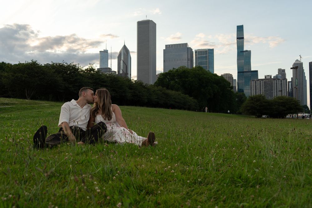 Couple sharing a kiss on green grass in Chicago's Grant Park with city skyline backdrop at dusk.