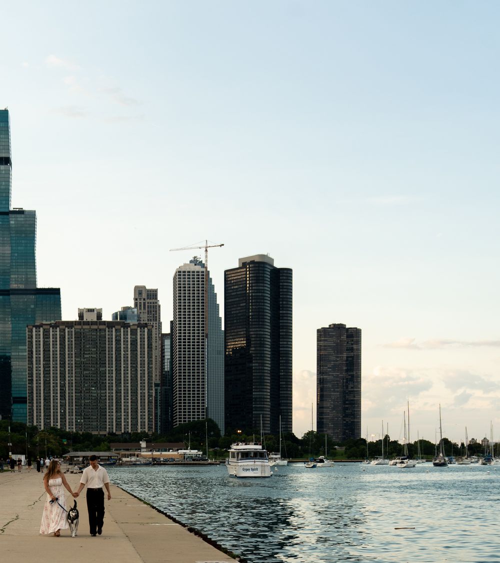 A couple in wedding attire walks along a lakeside promenade with the Chicago skyline and sailboats in the background.