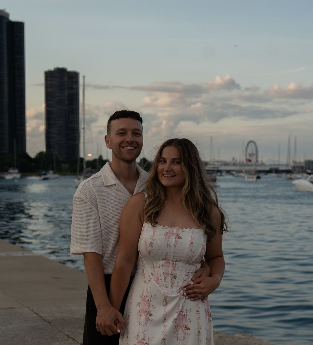 Couple smiling together on a waterfront promenade at dusk with city skyline and Ferris wheel in background.