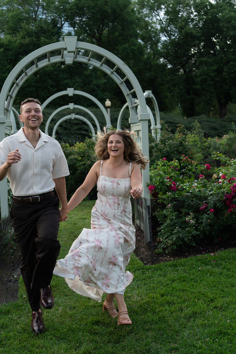 A joyful couple runs hand-in-hand through a lush garden with white arched trellises and colorful roses.