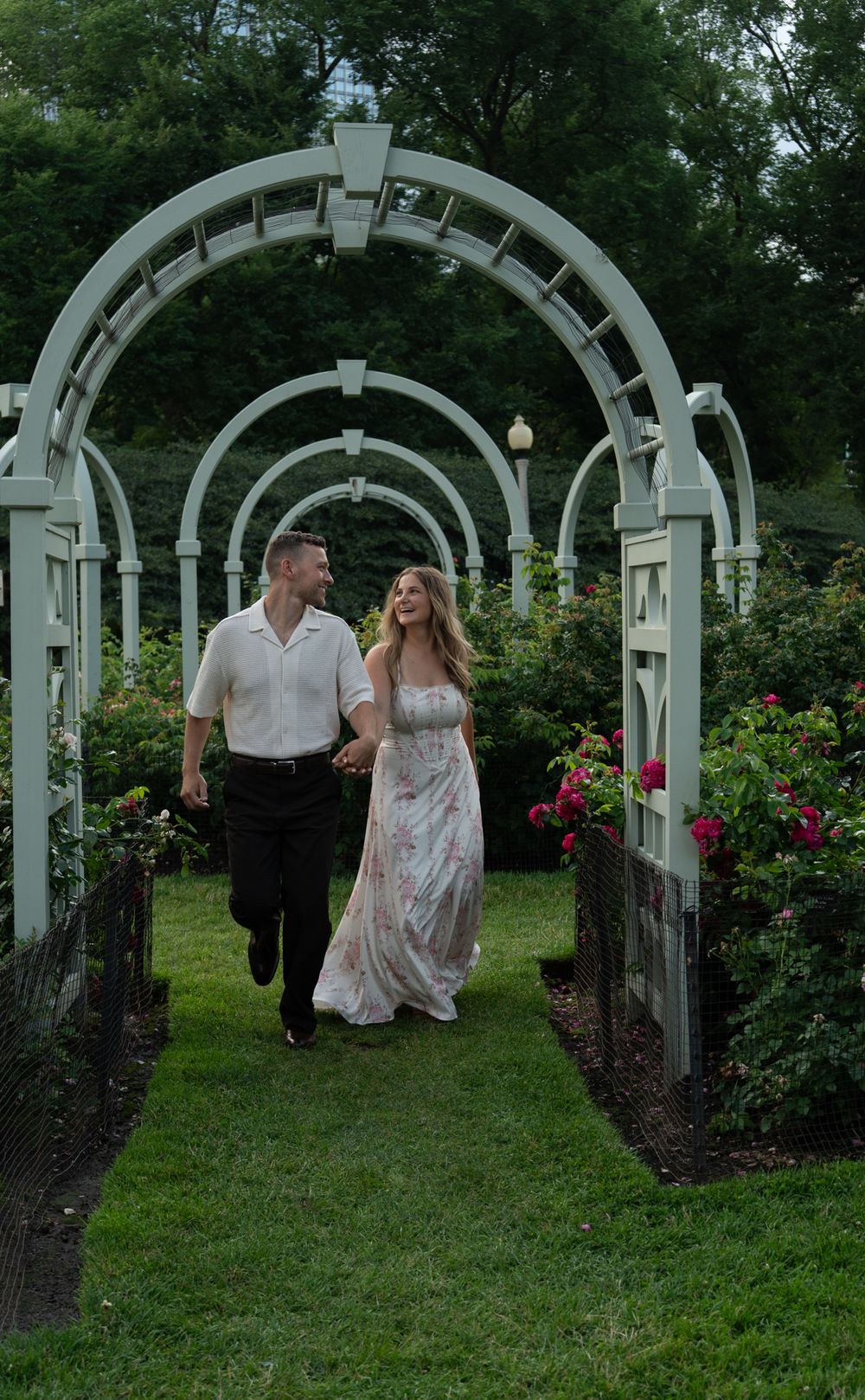 Couple holding hands walking through white garden arbor arches surrounded by blooming roses.