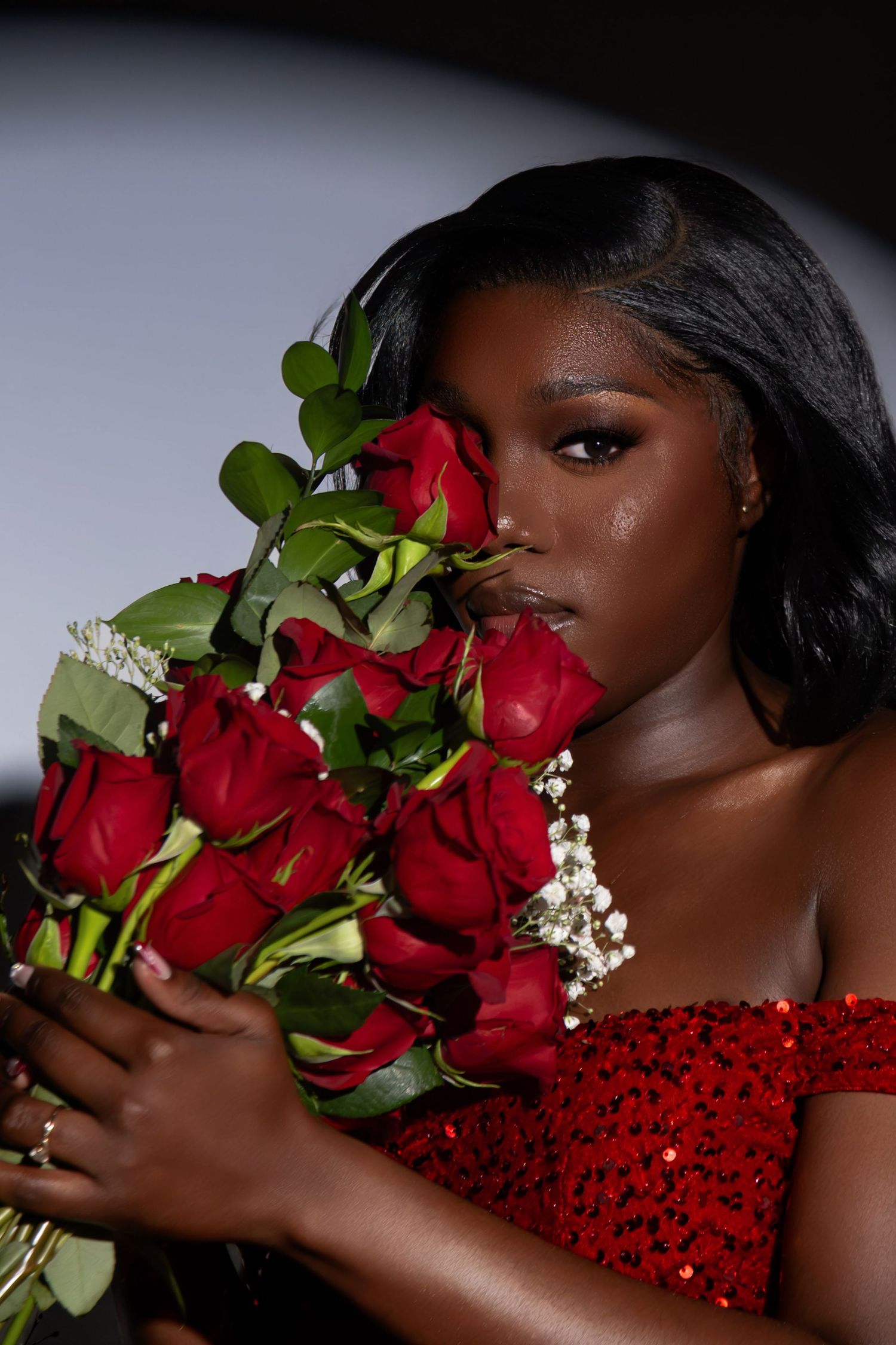 Woman in red sequin dress holding bouquet of red roses near her face in a dramatic studio portrait.