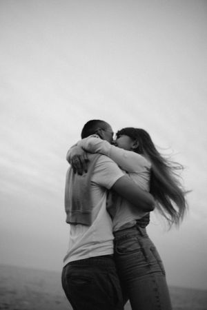 A couple embraces and kisses against a cloudy sky, with the woman's long hair flowing in the wind.