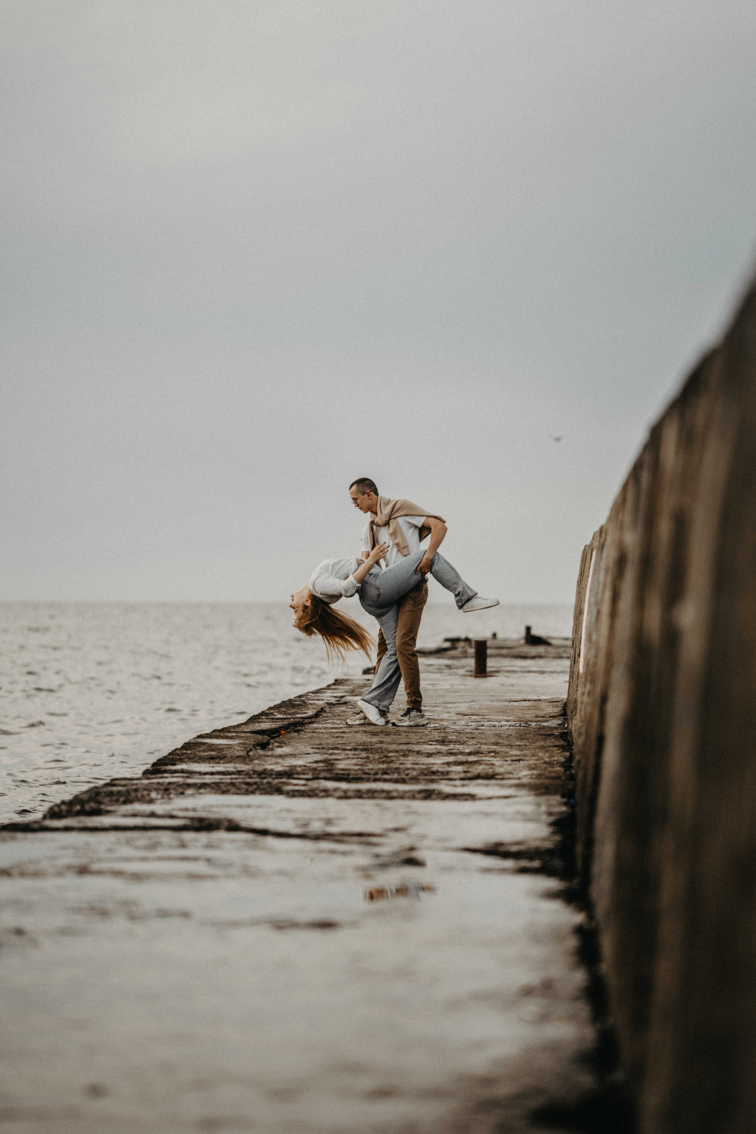 A couple dances playfully on a stone pier by the sea, with the man dipping the woman dramatically.