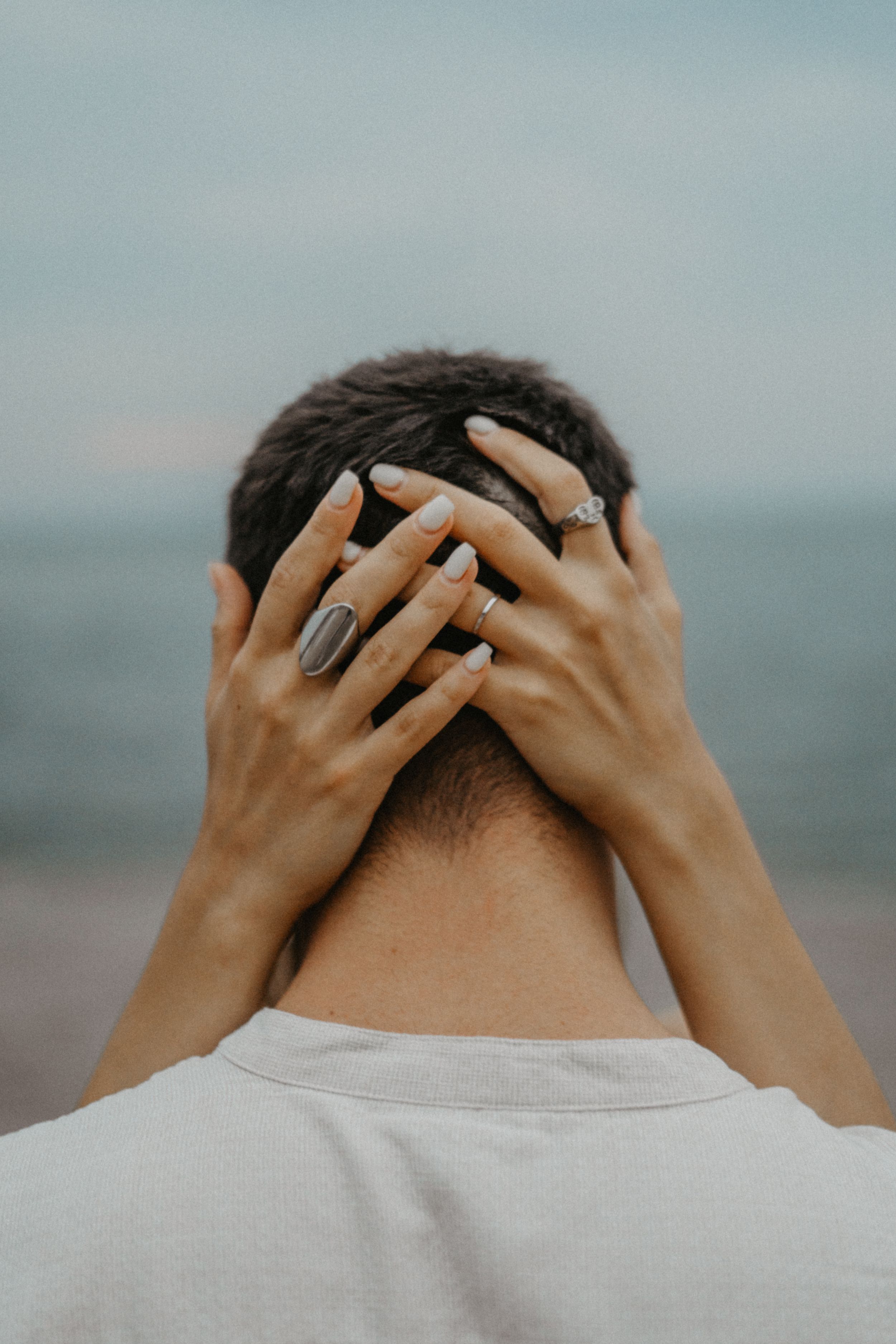 Woman with rings placing hands on mans head at beach.