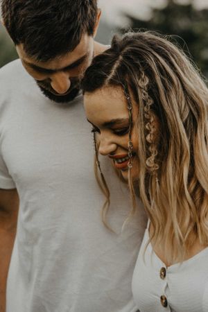 A smiling woman with braided hair accessories leans against a bearded man in a white t-shirt outdoors.