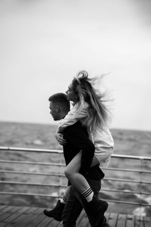 A man carries a woman on his back on a seaside pier in a playful black and white photo.