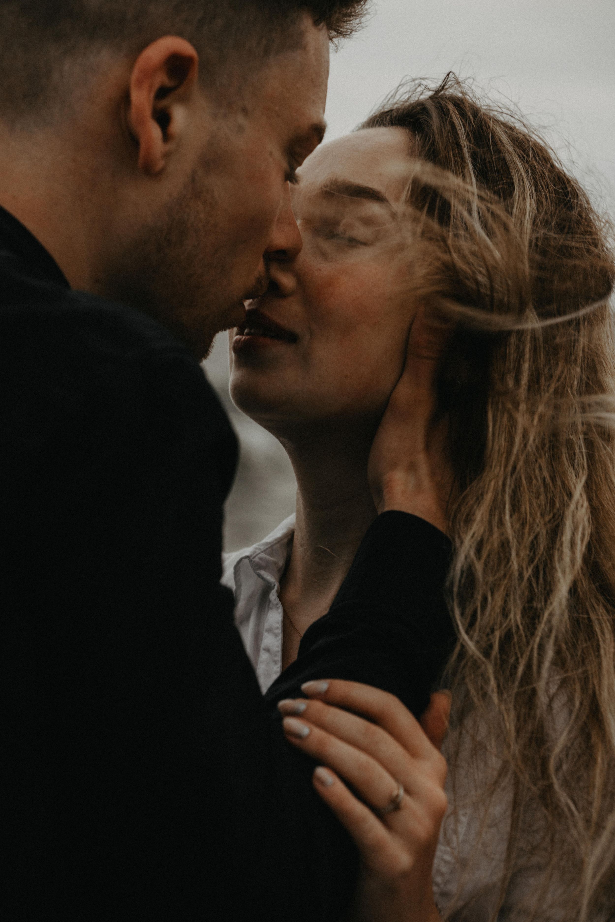 A couple shares a tender kiss outdoors, with the woman's hair blowing in the wind as he gently holds her face.