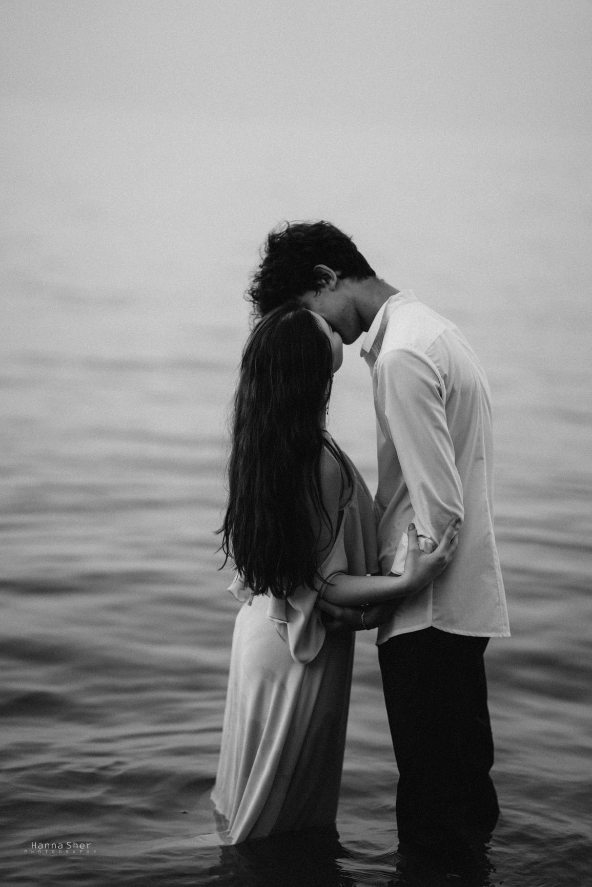 A romantic couple shares an intimate kiss while standing together in calm water, captured in black and white.