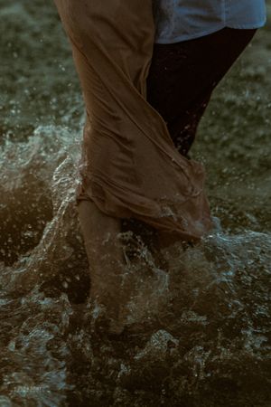 Close-up of a couple's legs wading through splashing ocean waves, with wet clothing and bare feet visible.
