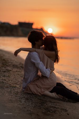 A couple sits closely together on a beach sharing a kiss, silhouetted against a stunning golden sunset.