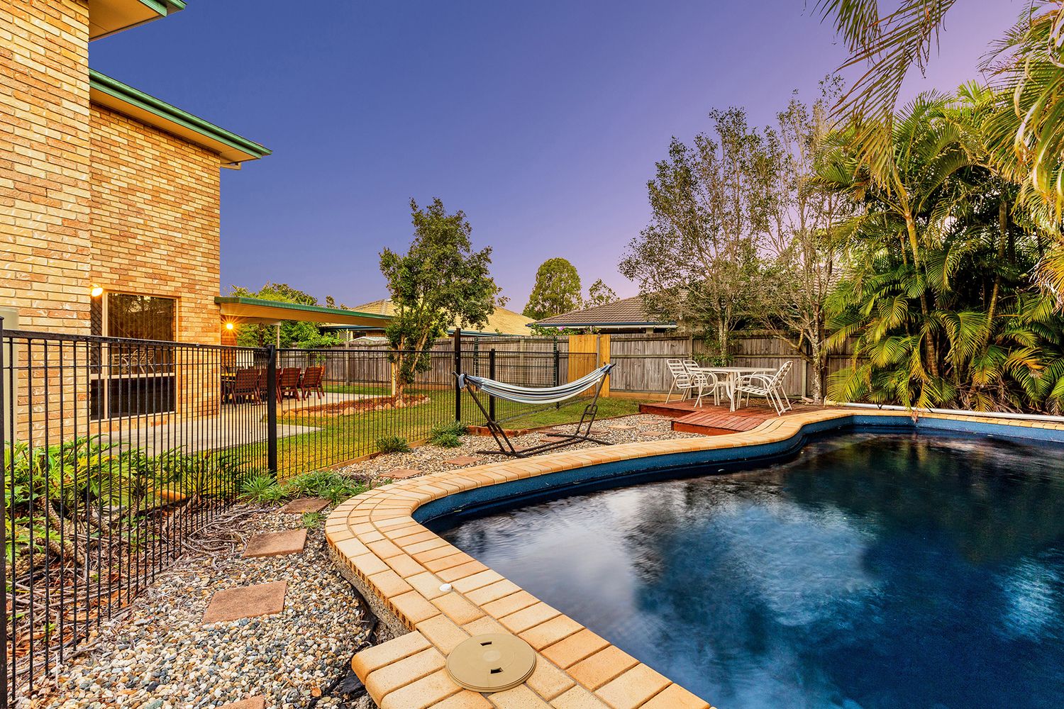 Backyard pool with brick surrounds, hammock, white outdoor furniture, and lush greenery.