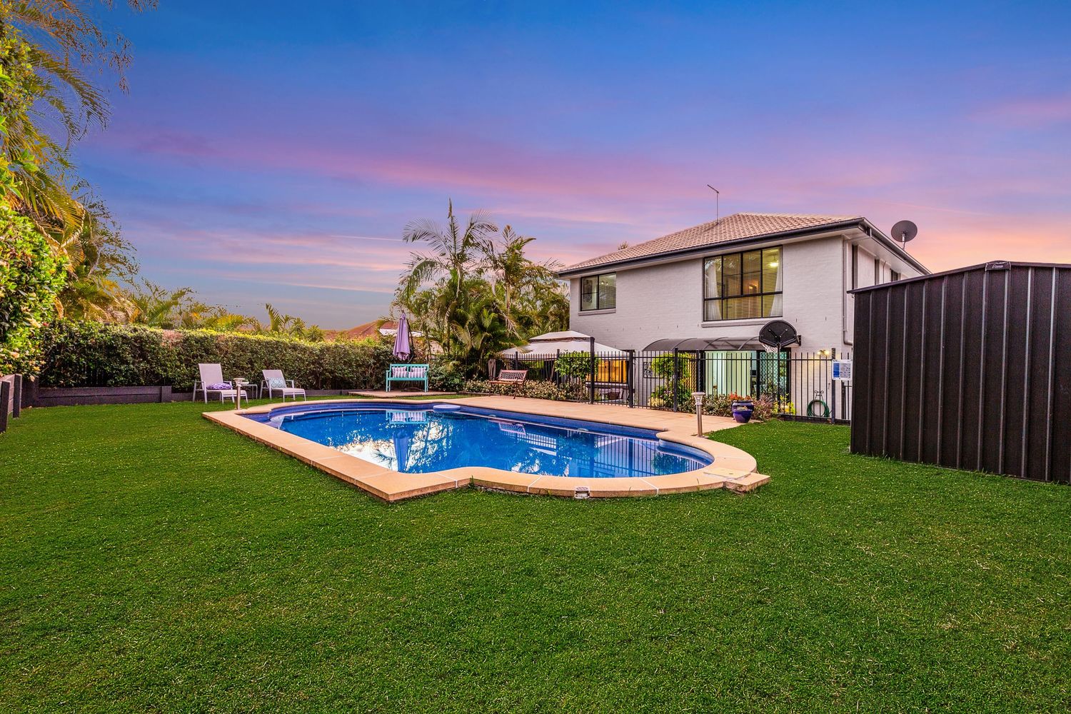 Backyard pool with kidney shape design, green lawn, patio chairs, and lush trees at sunset.