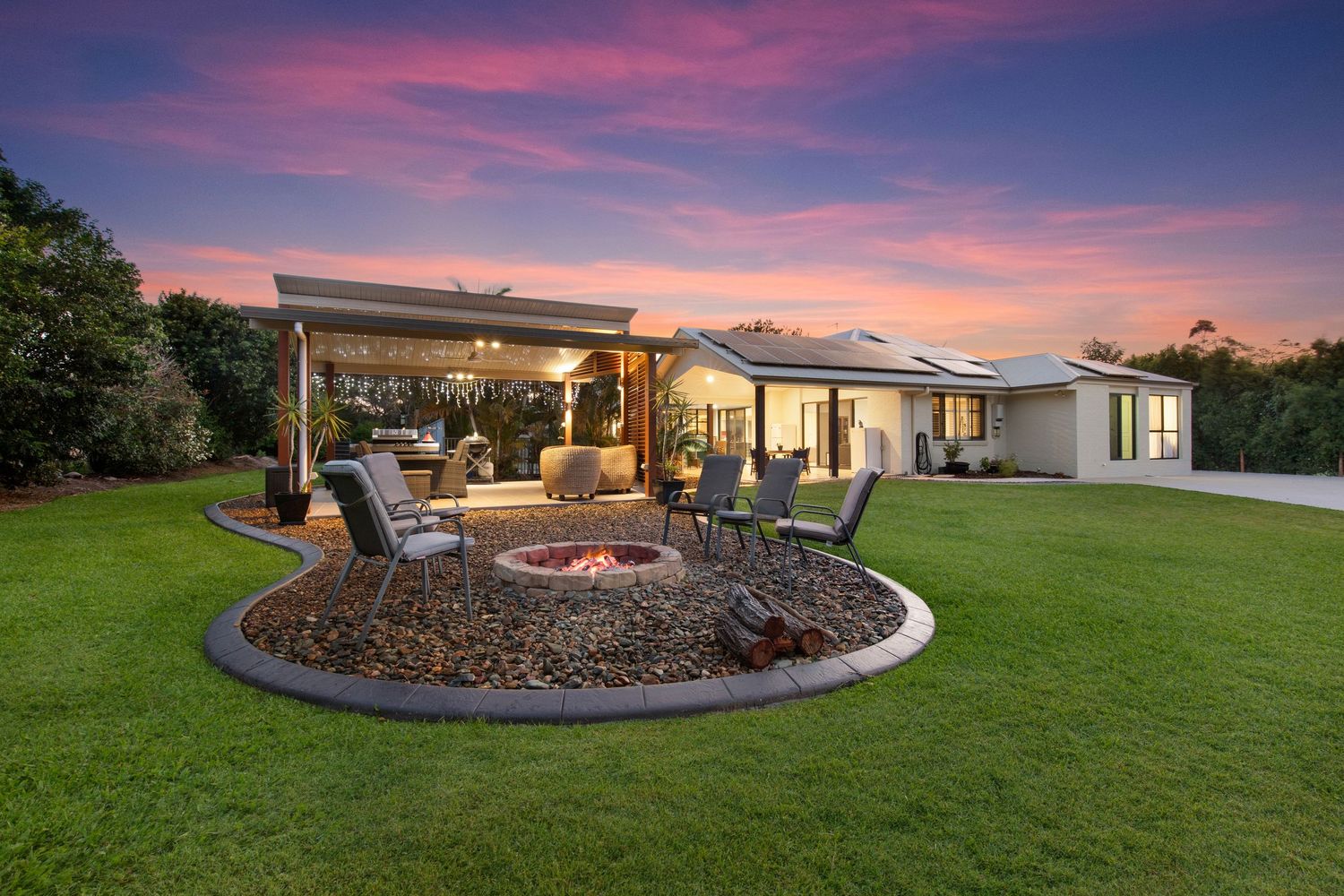 Large outdoor entertaining area with fire pit circle and pergola on manicured lawn at sunset.