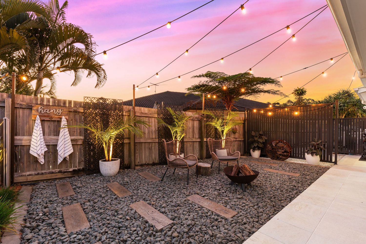 Charming backyard entertaining area with festoon lights, palm trees, fire pit, and decorative pebble garden at sunset.