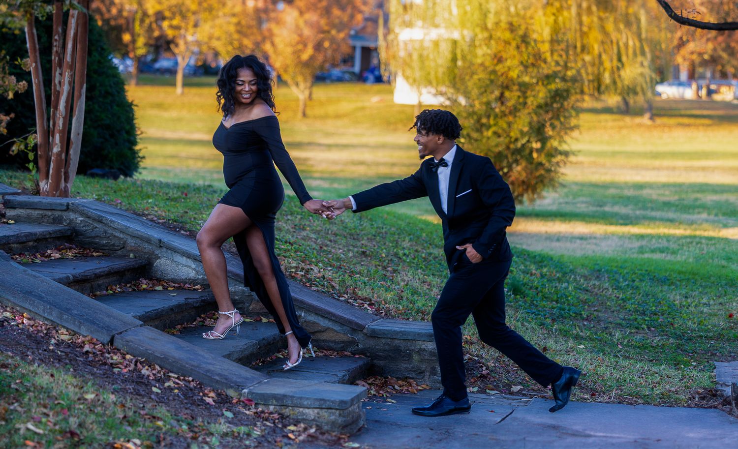 A couple in formal attire poses on stone steps in a park during autumn with golden foliage in the background.