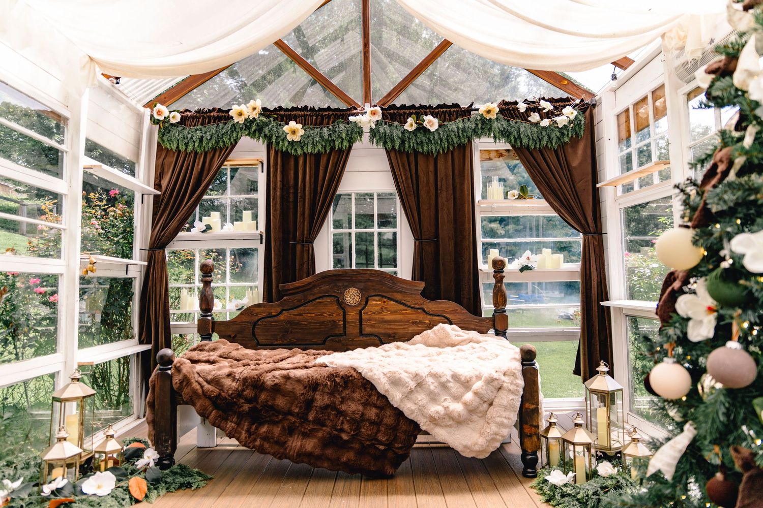 Rustic wooden bed with fur blankets inside a glass greenhouse styled for Christmas with a decorated tree, lanterns, and floral garland.