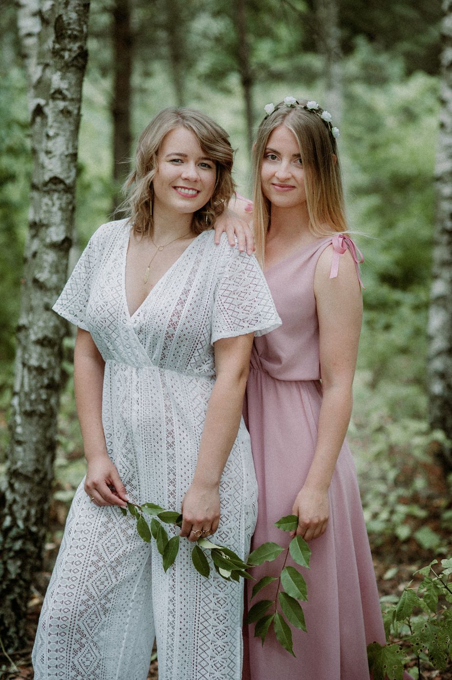 Two women in white lace and pink dresses pose together in a birch forest setting.