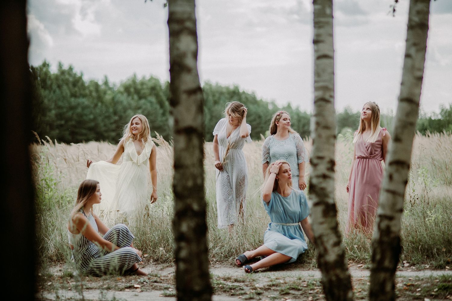 A group of people in casual outfits laugh and socialize together in a wooded outdoor setting during a photo session.