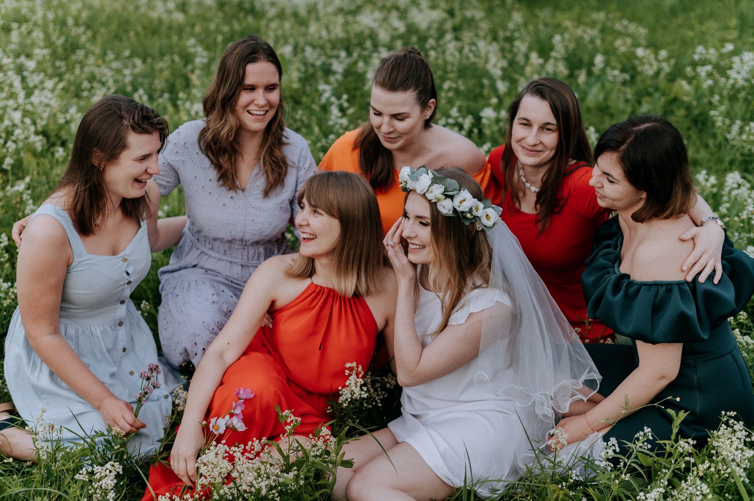 A group of friends in colorful dresses share joyful laughter while sitting together in a blooming wildflower meadow.