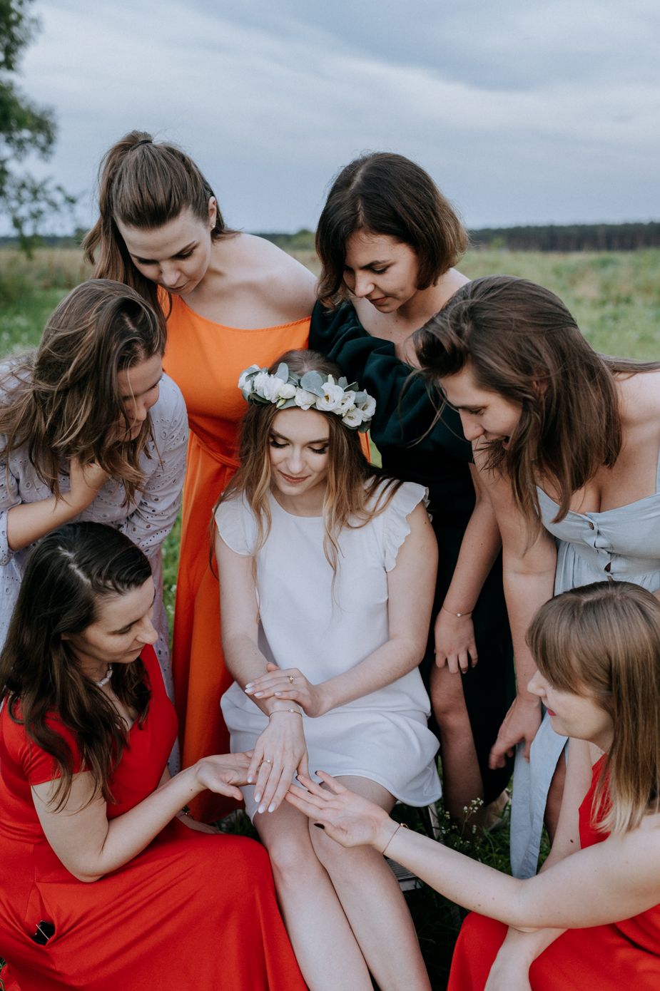 A group of friends in red and orange dresses gather in a circle outdoors examining something together.
