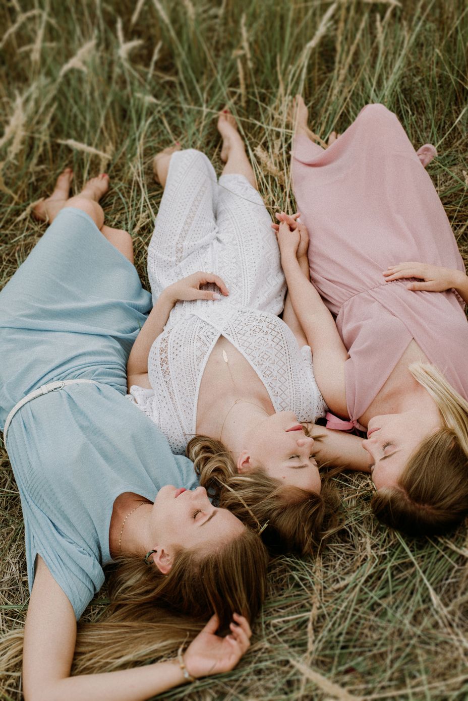 Friends lying in grass wearing pastel colors and white lace, creating a dreamy summer scene.