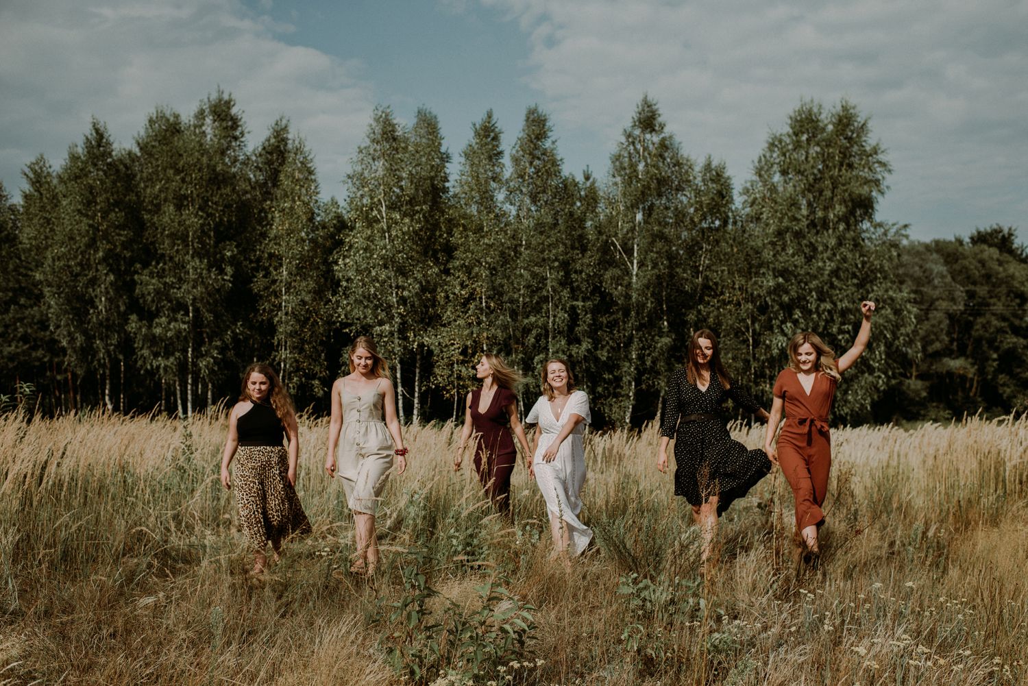 A group of people walk through a sunlit meadow with tall grass and trees in the background.
