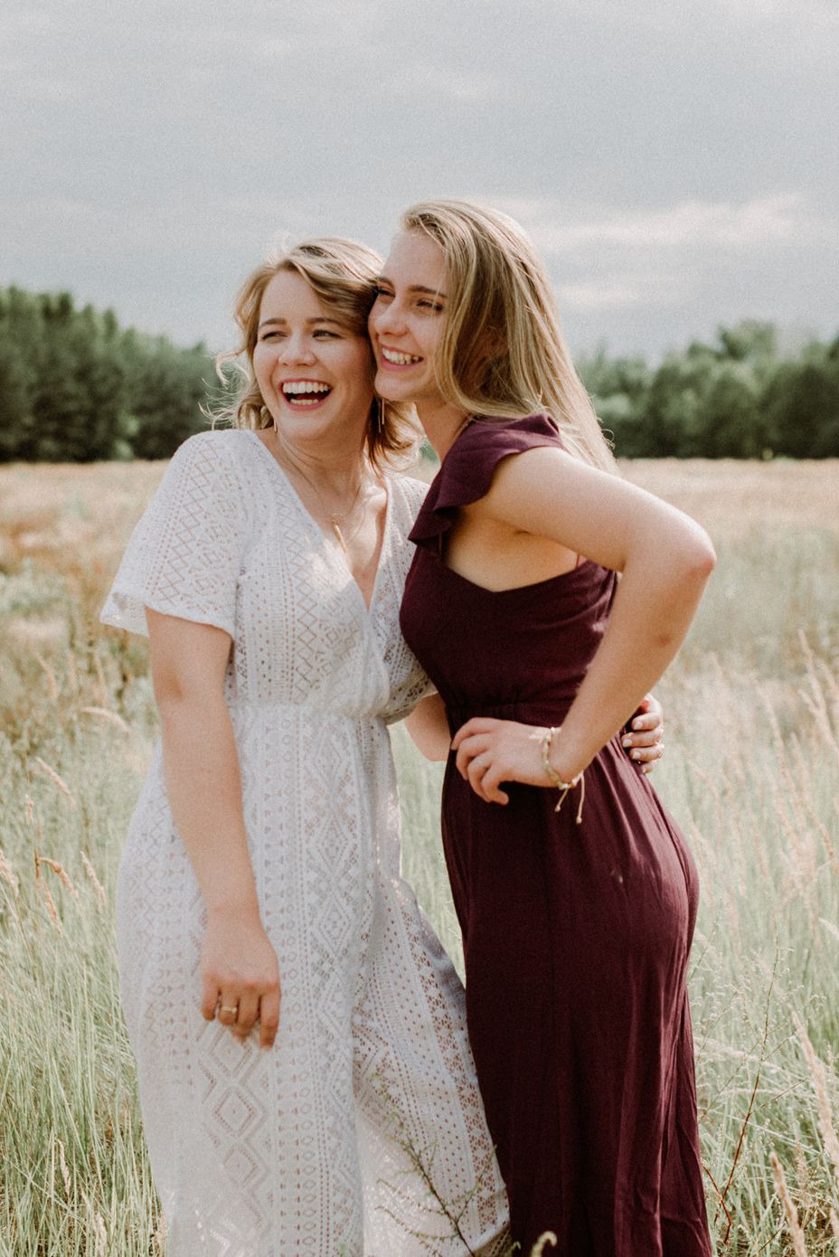 Two friends sharing a joyful moment together in a summer field wearing white lace and burgundy dresses.