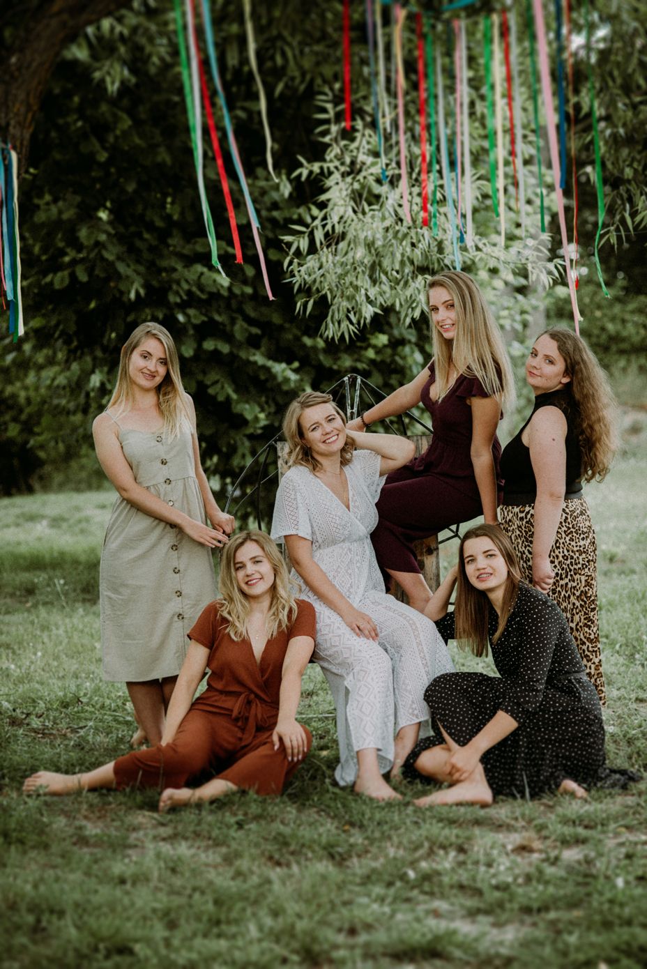A group of friends in casual dresses sit together on grass with colorful ribbons hanging above them in a dreamy outdoor setting.