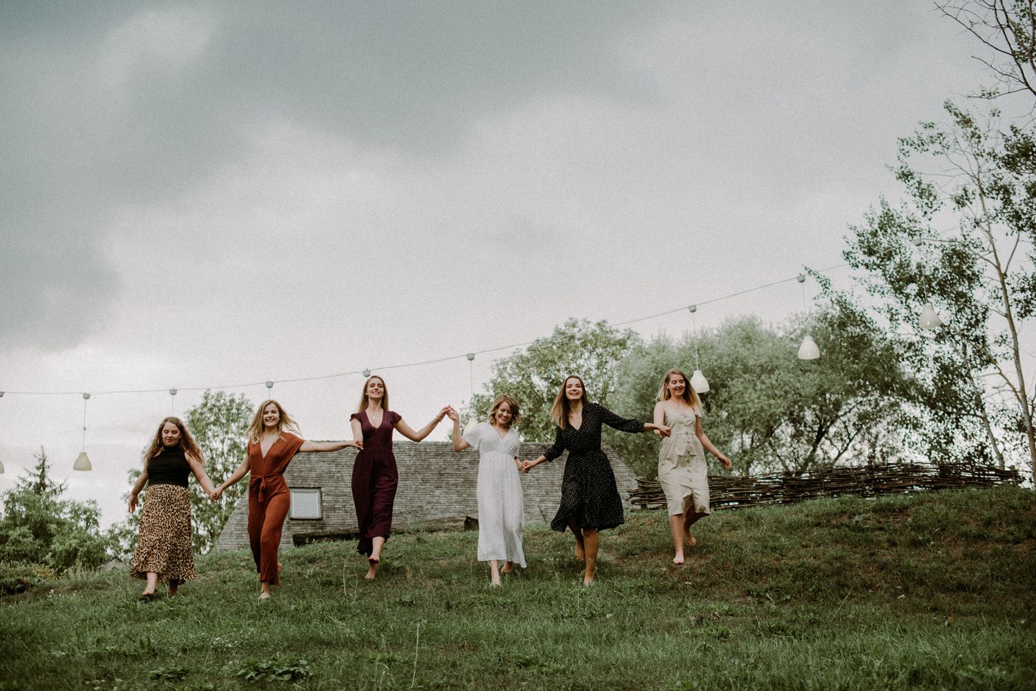 A group of people in flowing dresses run joyfully together on a grassy hilltop under moody storm clouds.