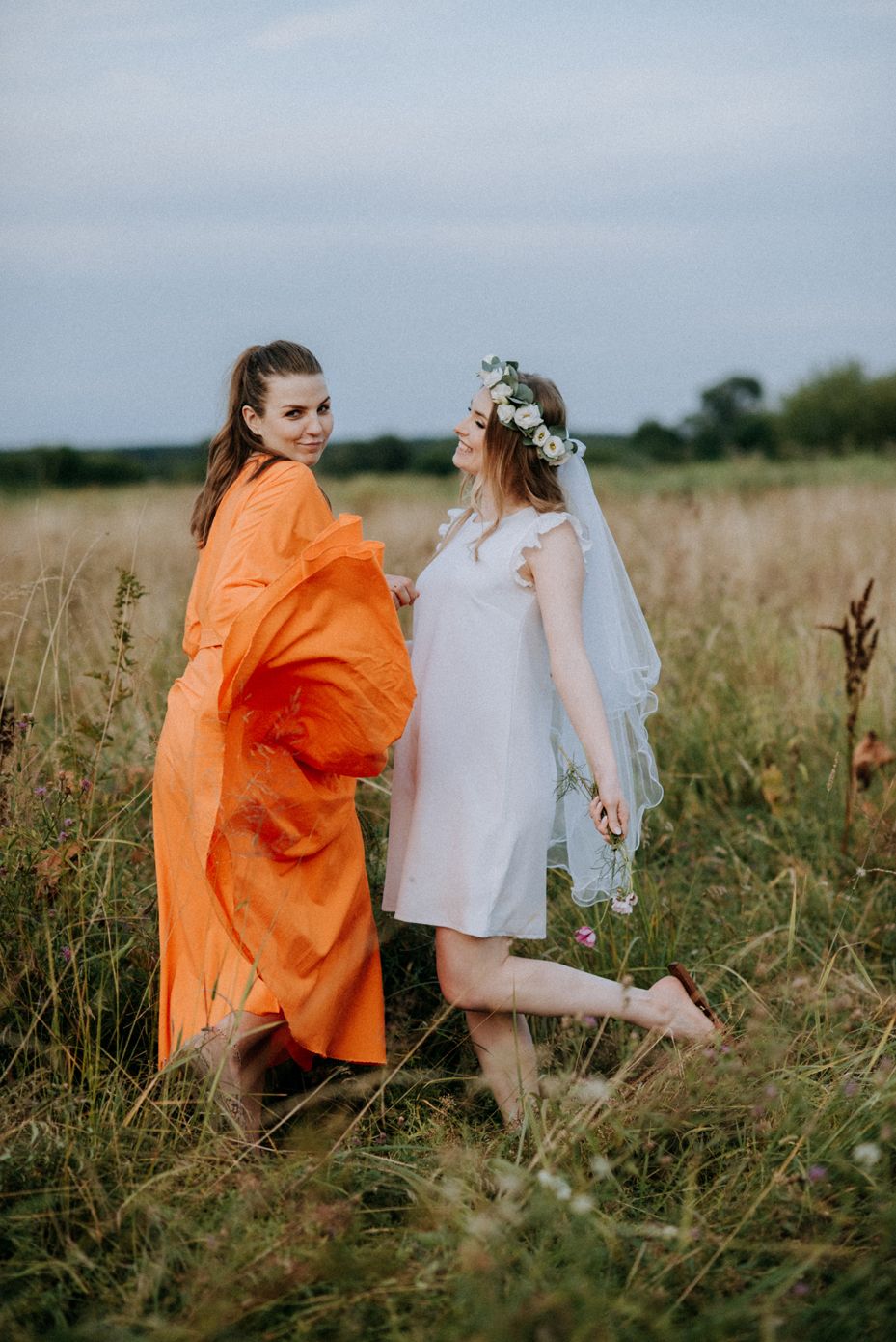 Two people in flowing dresses dance together in a summer meadow at dusk, one wearing orange and one in white with flowers.