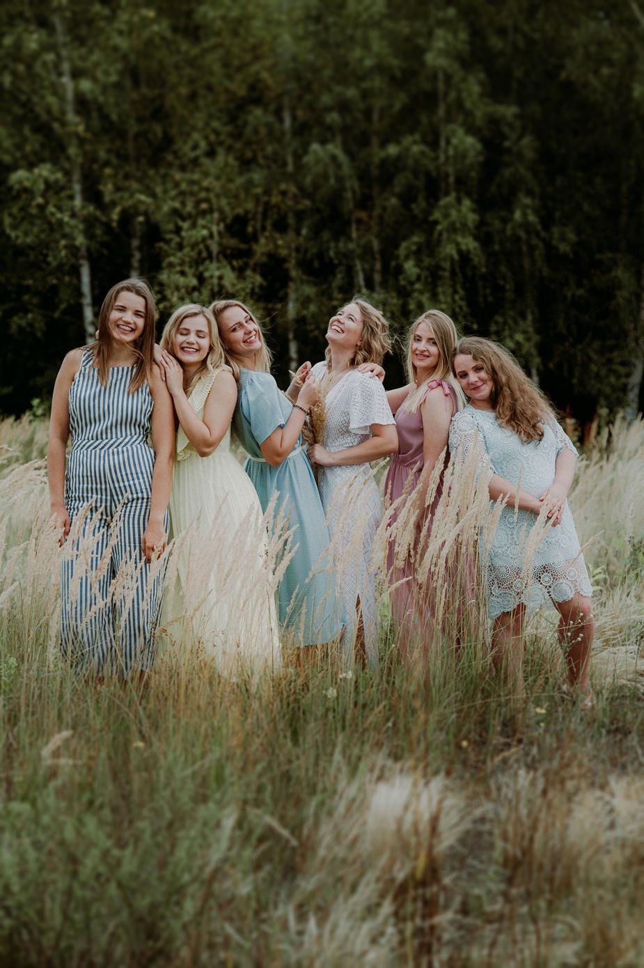 A group of friends in blue and white dresses laugh together while standing in a sunlit meadow of tall grass.
