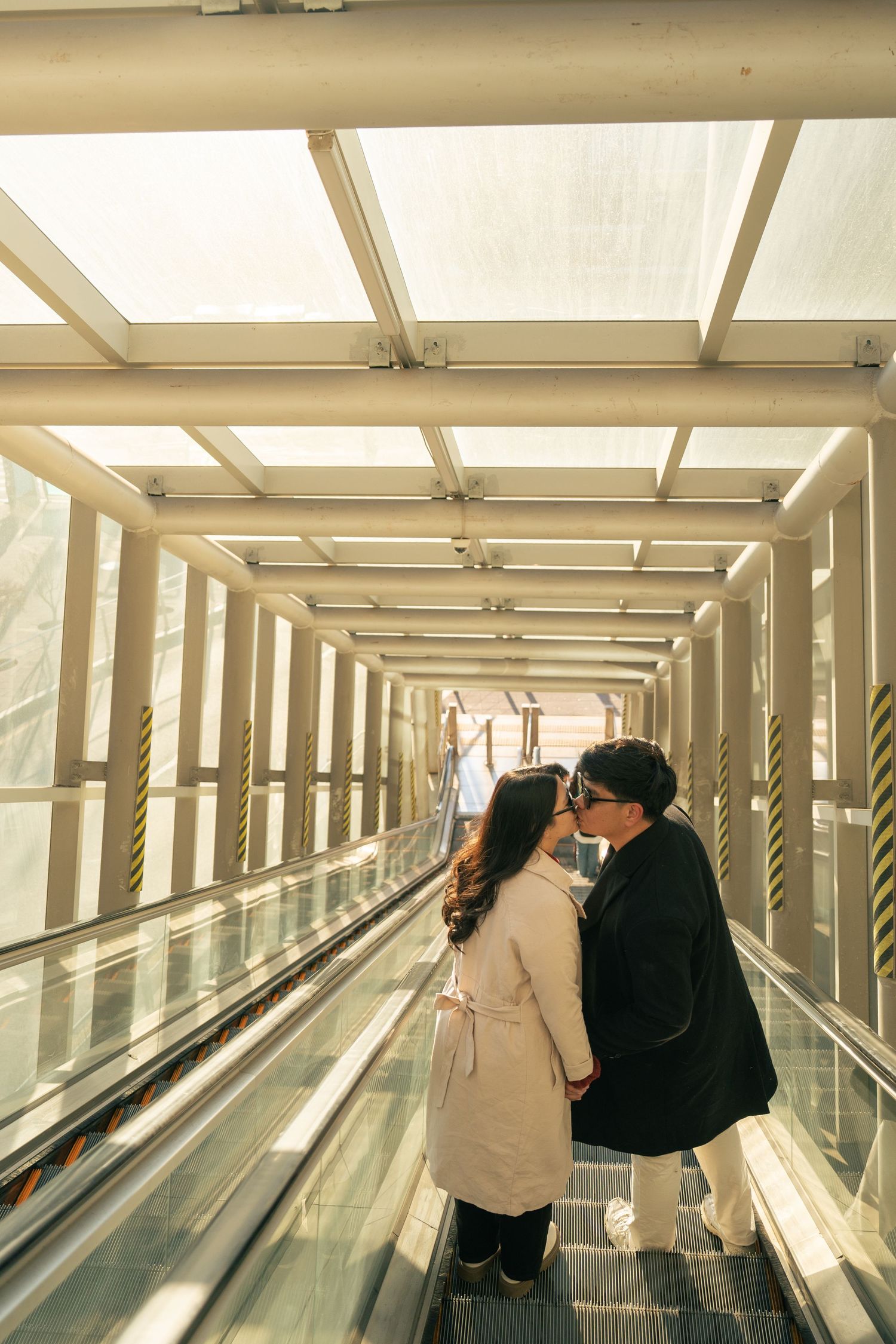 A couple stands close together on a moving walkway inside a modern glass-enclosed corridor with natural lighting.
