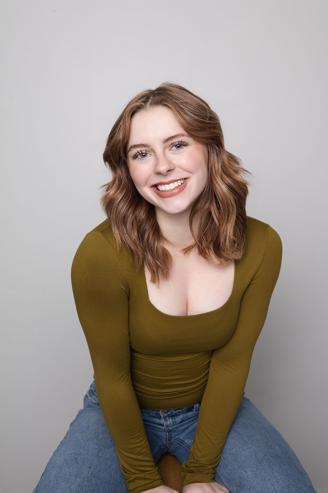 A person wearing an olive green long-sleeved top and jeans poses against a gray background with a warm smile.