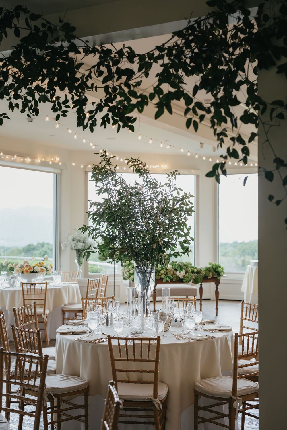 Elegant wedding reception venue with white tables, wooden chairs, and natural light streaming through large windows.