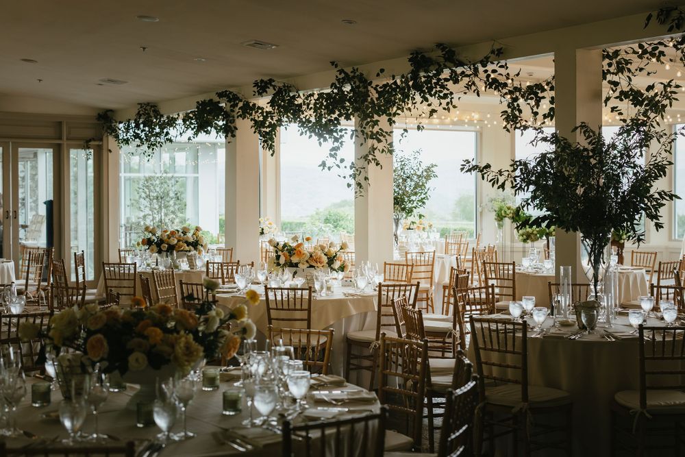 Elegant wedding reception venue with white tablecloths and chiavari chairs decorated with floral arrangements and natural light.
