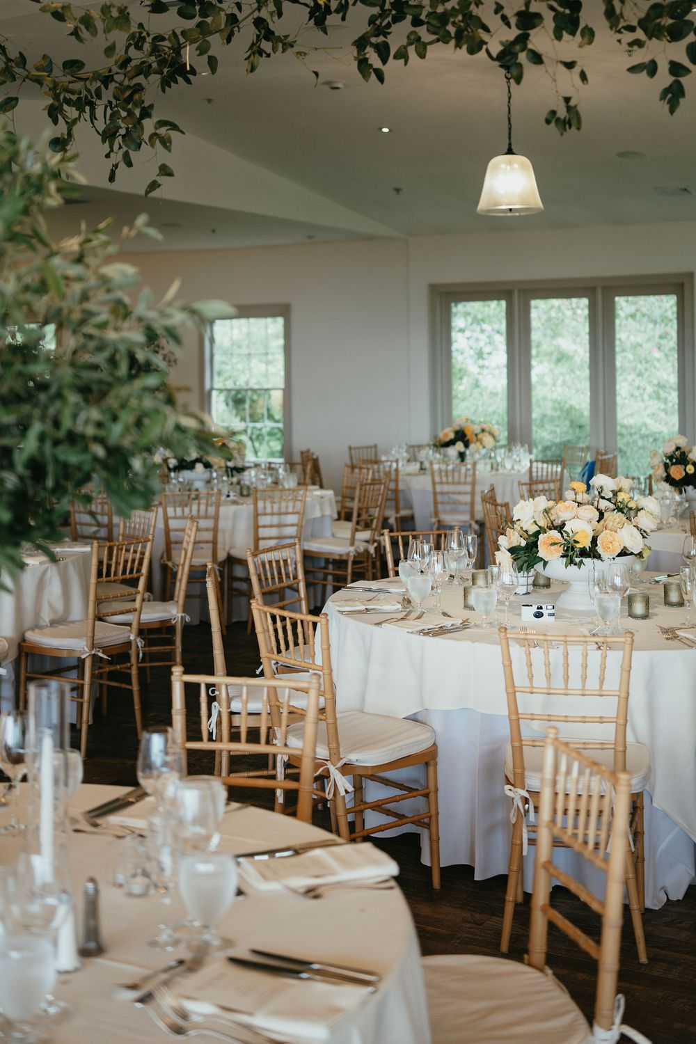 Bright dining room setup with round tables, chiavari chairs, and greenery decor hanging from above.